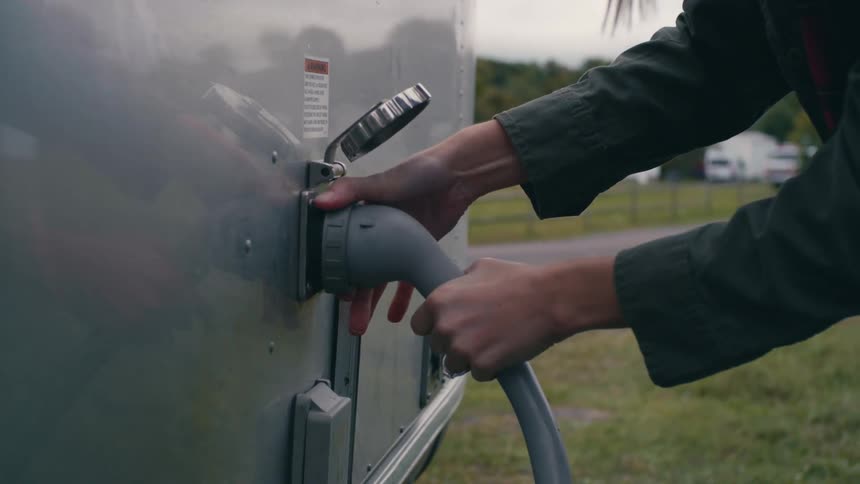 Woman wearing a sun hat and a coat fixing a pipe with a cord on the back of a silver caravan