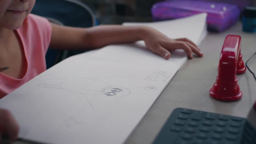 A girl sitting at a desk drawing on a paper with a pencil