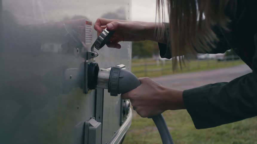 A woman with a hat attaching a hose to a vehicle with trees the sky and clouds in the background