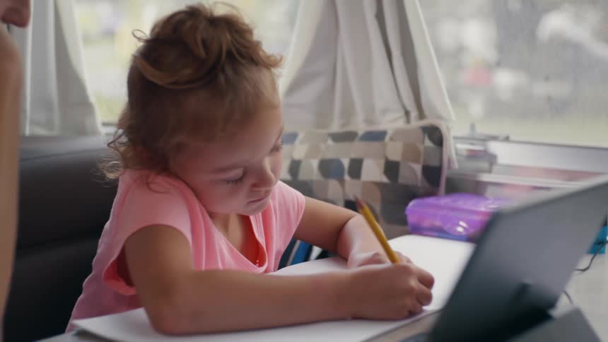 Mother and daughter sitting inside of a home at a table in front of a laptop while the daughter works on school work