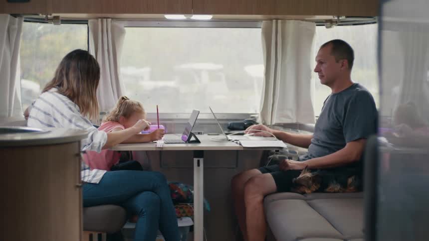 A young girl draws as her parents work on their laptops in the camper