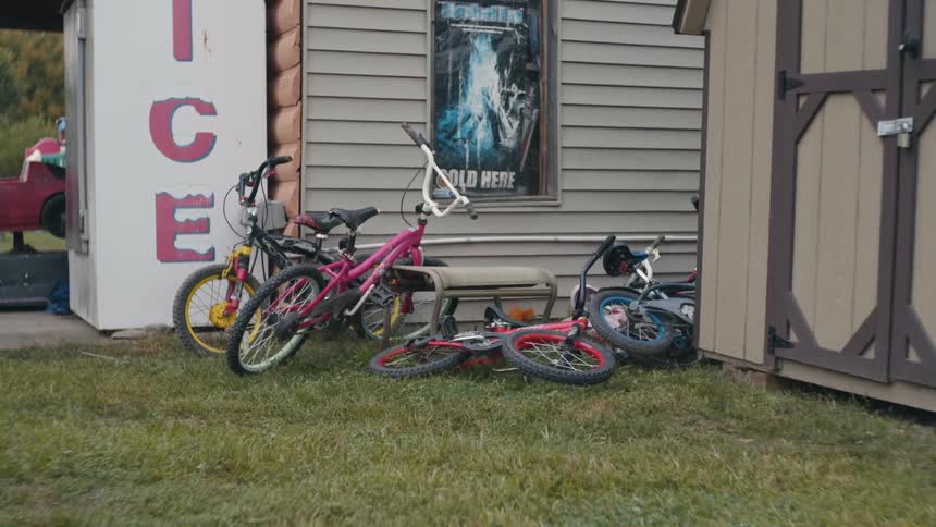 A set of bikes lying around in the grass outside of a backyard next to a brown shed at daytime