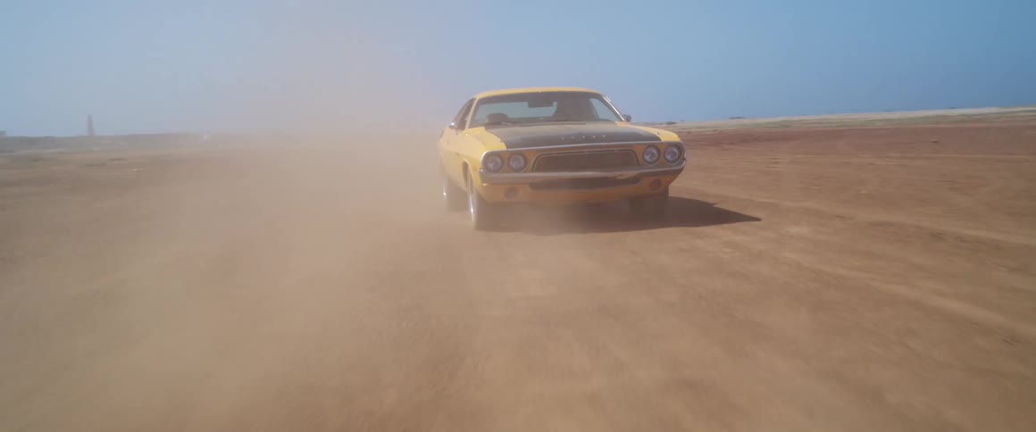 Car drives down dusty dirt road or track with the windows down and a building in the background on a sunny day with a bright blue sky