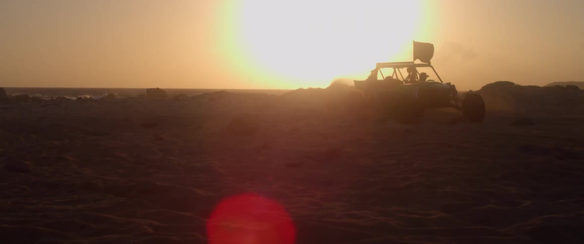 Atv with a flag above it drifting through sand in front of a sunset