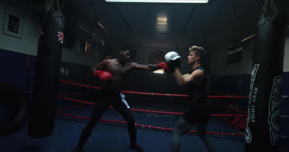 Man wearing boxing gloves inside of a boxing ring indoors practicing with another man wearing protective mits