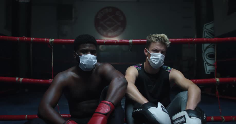 Two male boxers with white masks sits down in the boxing ring side by side