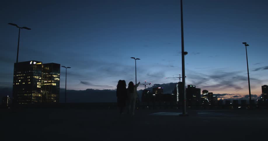 Two people walk at night with light poles above them that are off with big buildings and a skyline in the background