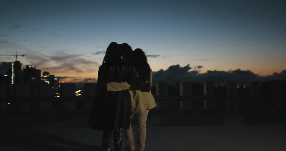 Two women holding each other walking on a sidewalk next to a bridge overlooking a city skyline