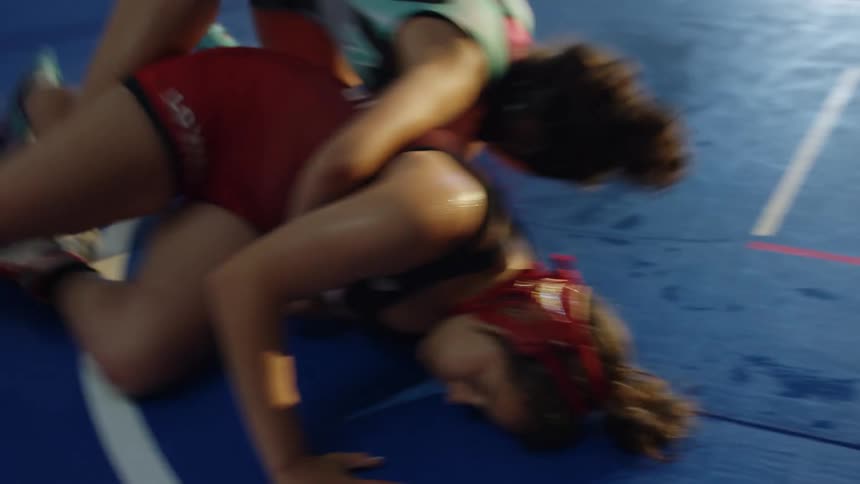 Two women are wrestling on a blue mat with white tape indoors
