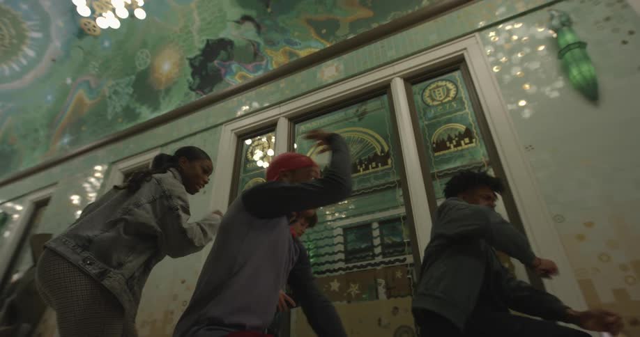 Teenagers dance in a corridor of an ornately decorated interior of a building with painted ceilings and unique green glass sconces