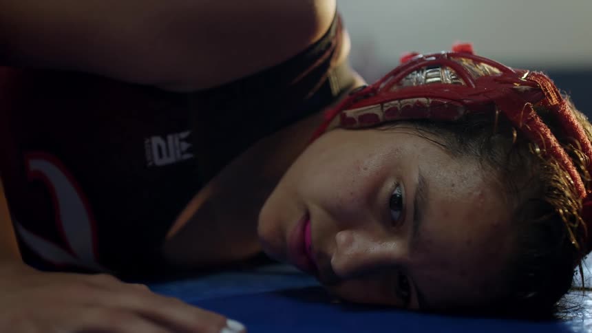 A woman with headgear and a mouthguard falling on a mat in a gym