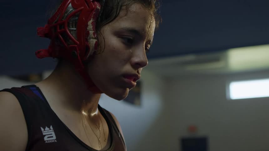 Teenage girl staring down wearing wrestling gear covered in sweat inside of a building