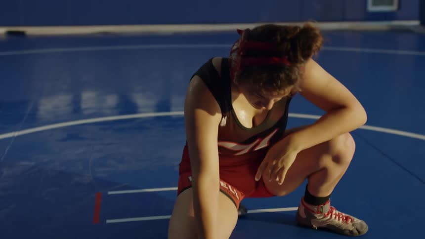 Teenage girl taking a breather inside of a blue gym at wrestling practice while another girl walks by