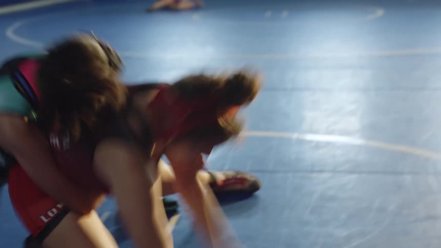 Two women wrestle on a blue mat in a gym during the day with two men in the background