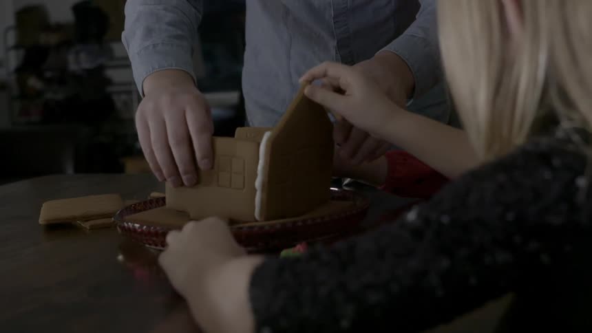 A man and a young girl assemble a gingerbread house
