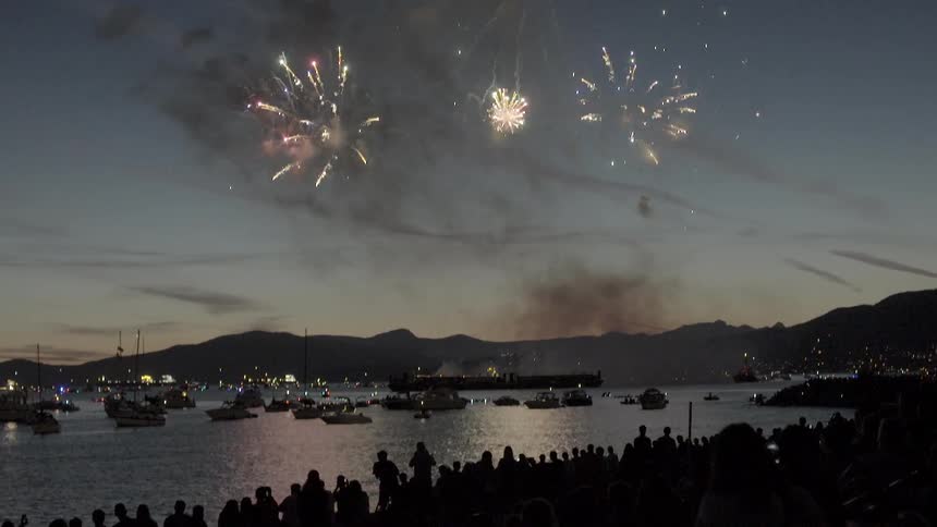 Fireworks explode over a harbor in a light night sky