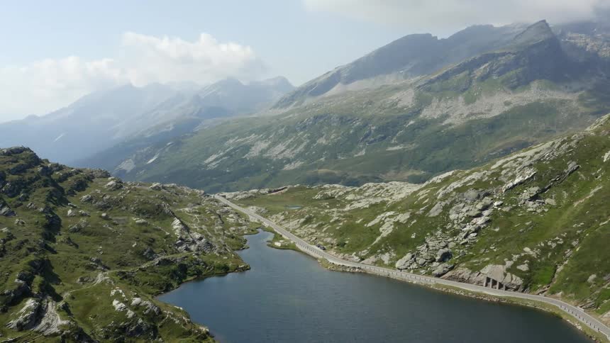 A lake in a mountain valley beside a winding road with fog in the distance on a sunny day