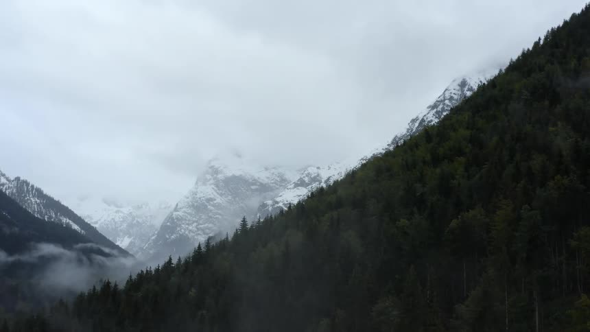 A mountain covered in evergreen forest surrounded by snow capped stone mountains on a cloudy day