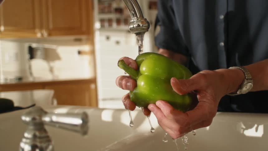 A man washing a green bell pepper under a kitchen faucet
