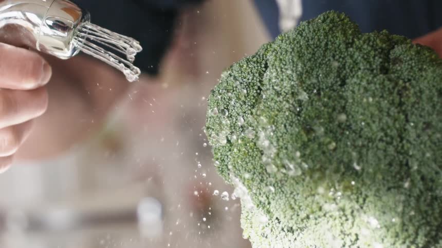 A person holds a head of broccoli in one hand and uses a sprayer to wash the broccoli with water in the other