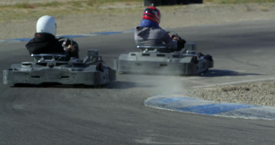 Two people with helmets racing go karts on a track with the sky and clouds in the background
