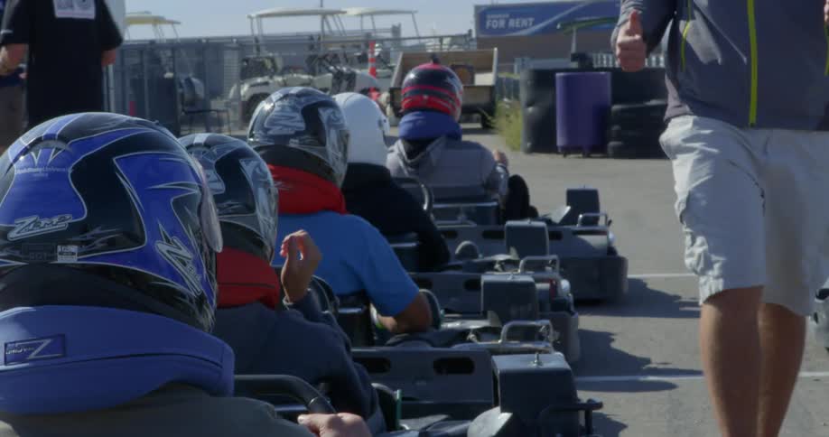 People sitting at a start line in go karts wearing helmets while a man does a safety check