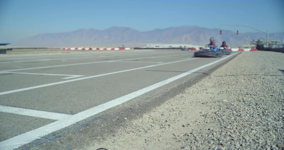 People with helmets racing go karts on a track with a mountain range the sky and clouds in the background