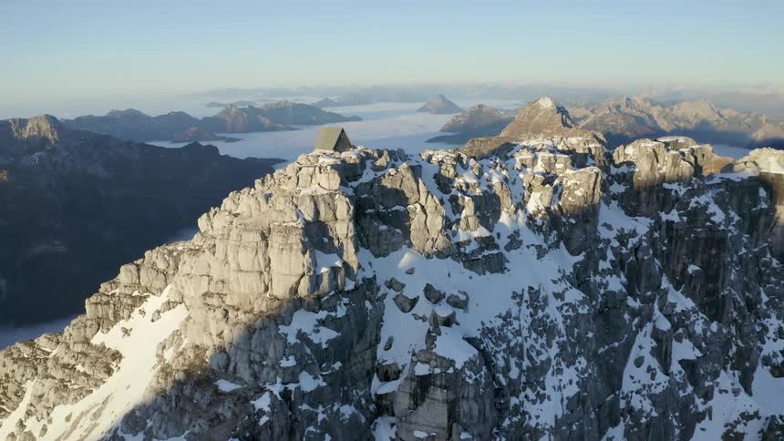 A cabin on a cliff with snow and ice with a mountain range the sky and clouds in the background