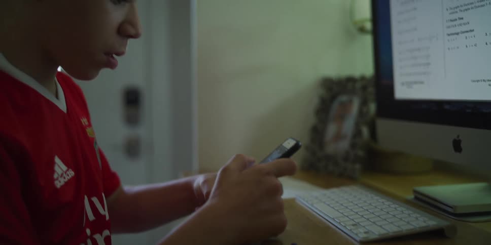 Teenage boy sitting at a desk in front of a desktop computer texting on his phone
