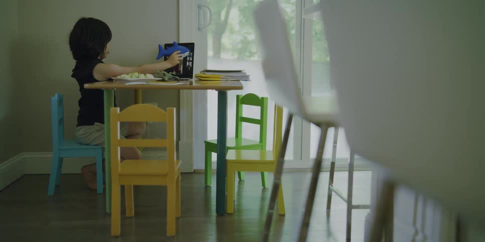Child playing with a toy shark pushing it against a screen of a tablet on a kids table indoors