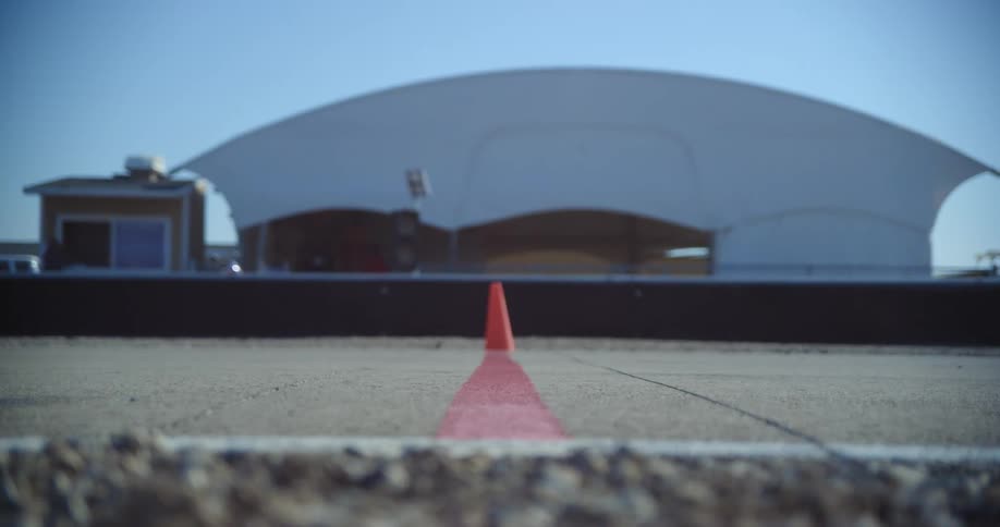 Go carts racing on a track with a cone and a red line with a building the sky and clouds in the background
