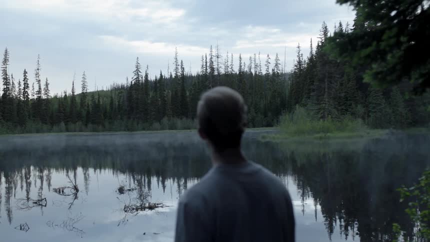 A man is looking over a lake towards a forest and hillside