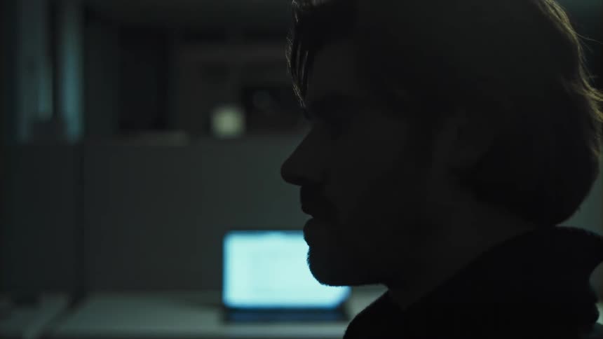 A man sits motionlessly in a cubicle within a darkened office space