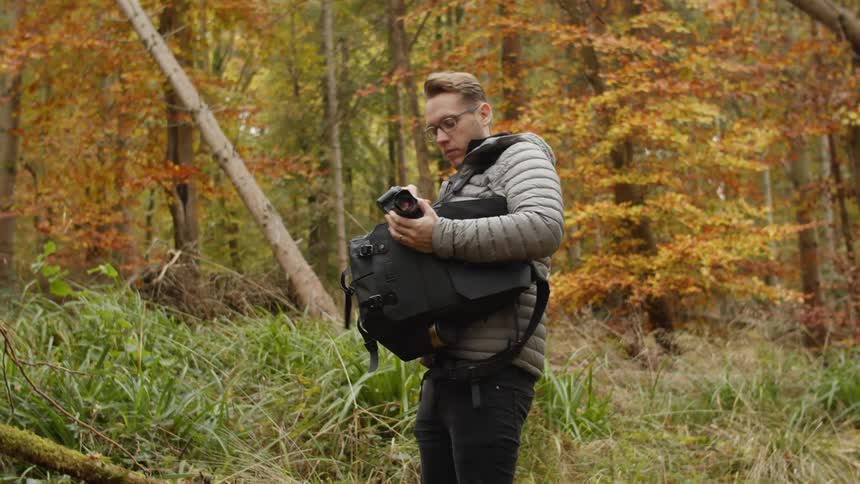 A photographer stops in a forest to put his camera in his black backpack