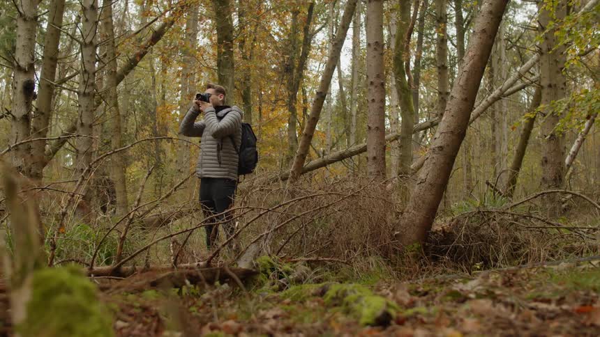 Male photographer with a grey coat and a black backpack take pictures in the woods