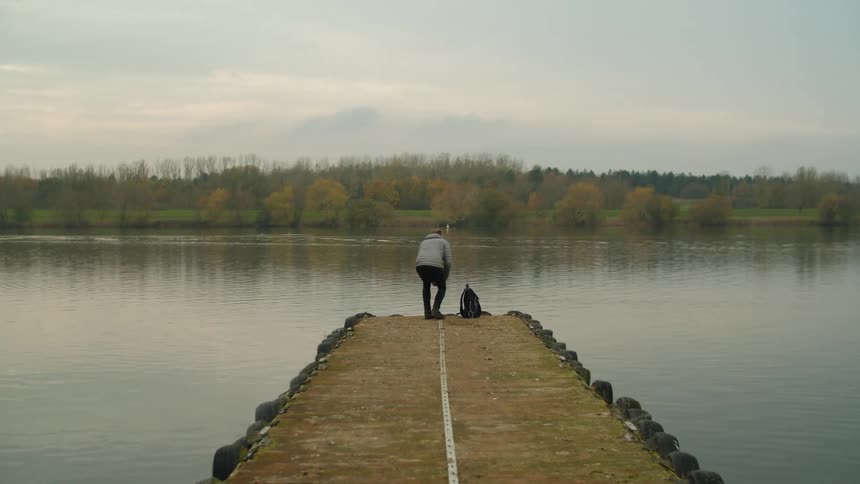 Man getting up from a squat standing at the edge of a pier looking out at a lake