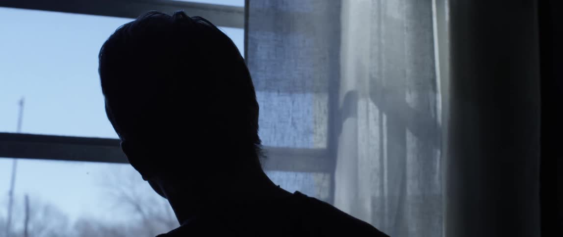A man stands inside without lights on looking out of a window