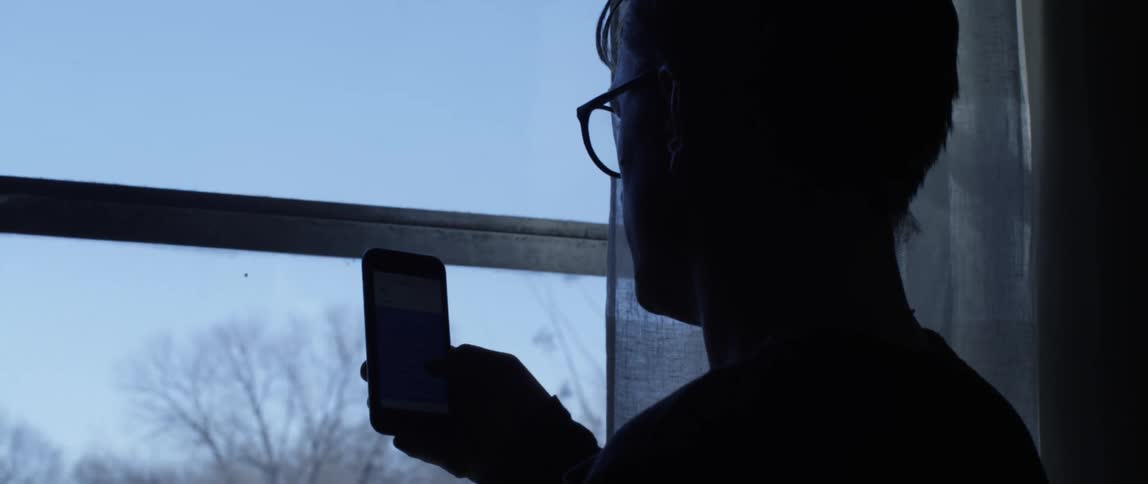 Man holding up and looking at a smartphone in front of a window inside of a room