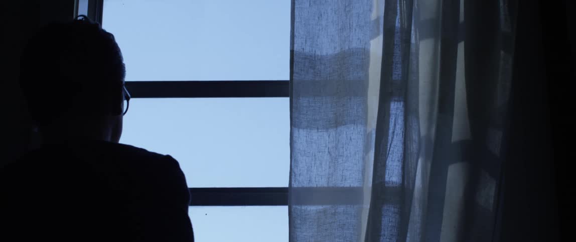 A teenager wearing glasses walks up to his window to look outside