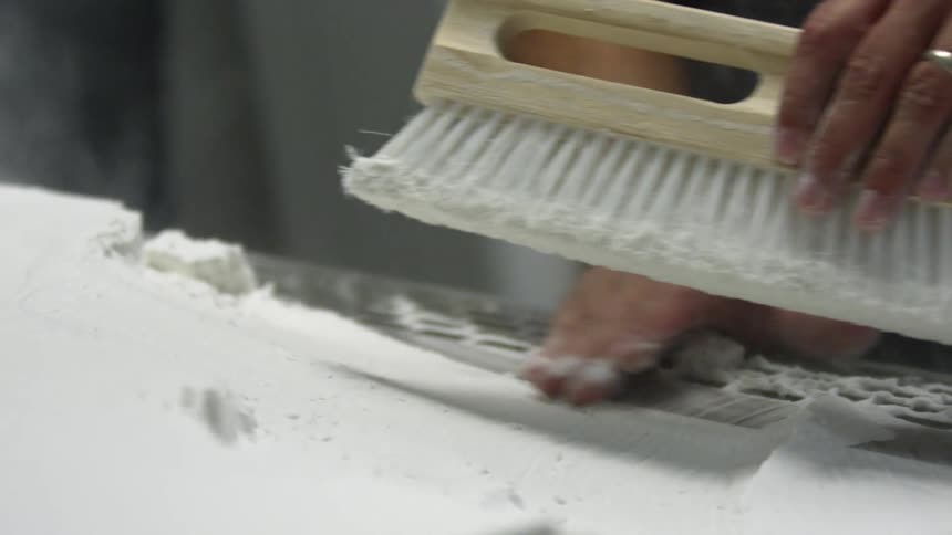 A man uses a brush to move and smooth white powder on a surface with a metal grate