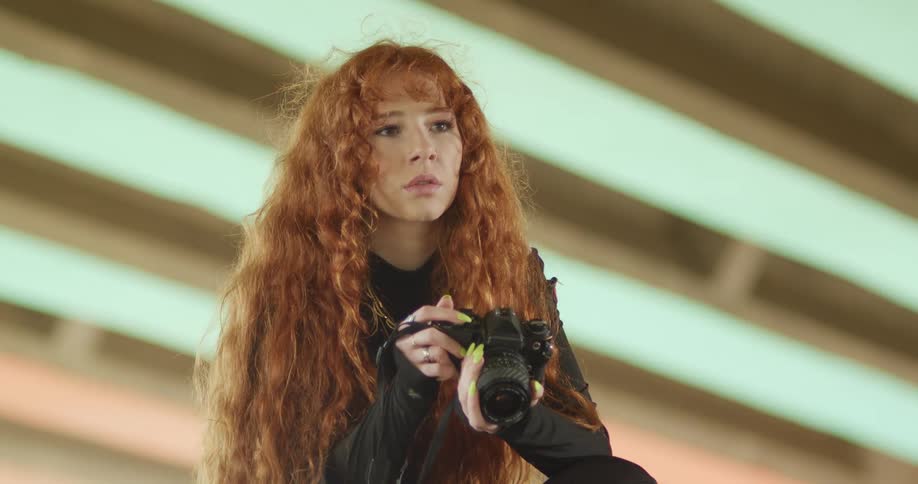 A woman with long red hair sits and takes a photo with her camera indoors