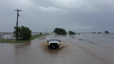 A utility vehicle driving on a flooded road with flooded farm land and heavy rain clouds in the background