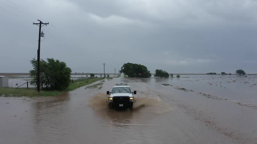 A utility vehicle driving on a flooded road with flooded farm land and heavy rain clouds in the background