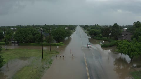 Aerial view of a vehicle driving over a flooded street on a small town while small kids play in the flooded roadway