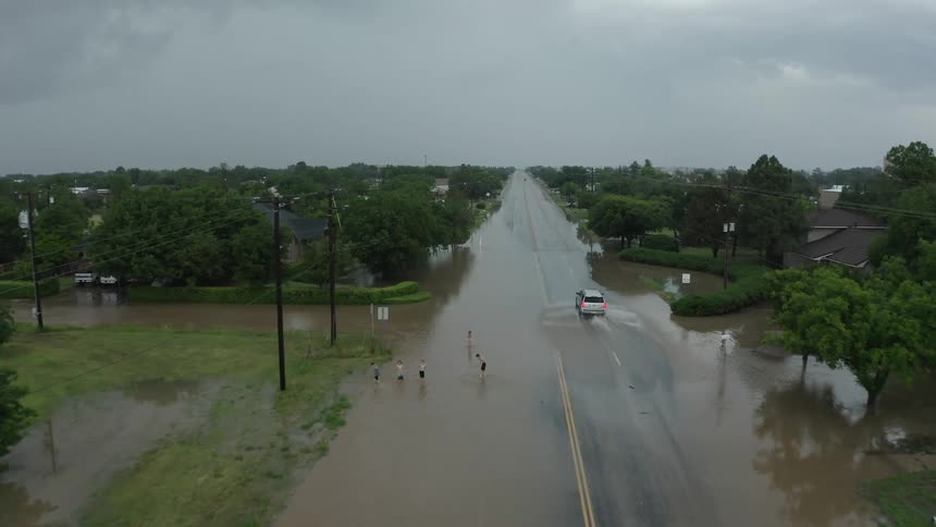 Aerial view of a vehicle driving over a flooded street on a small town while small kids play in the flooded roadway