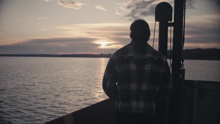 A man standing on a boat traveling on a lake looks out at the horizon in the evening