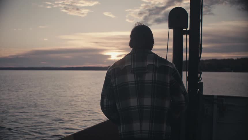 A man on a boat looks out at the sunset and then turns and walks away