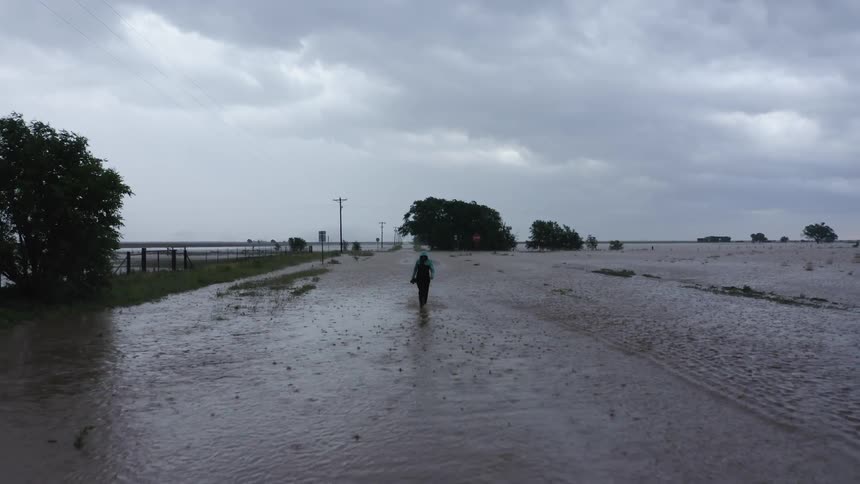 Aerial shot of a woman walking through flood water and the rain as she navigates a flooded road under heavy rain clouds