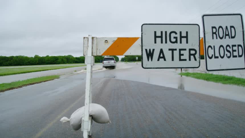 A high water and road closed sign is shown in the foreground as a white SUV drives across the flooded roadway