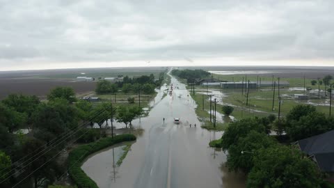 A car leaves a wake as it travels on a flooded road while small children play in the splashing water